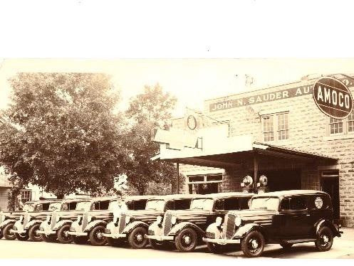 A row of vintage cars lined up in front of a automotive service station with an 'AMOCO' sign and 'John N. Sauder Auto' visible on the building