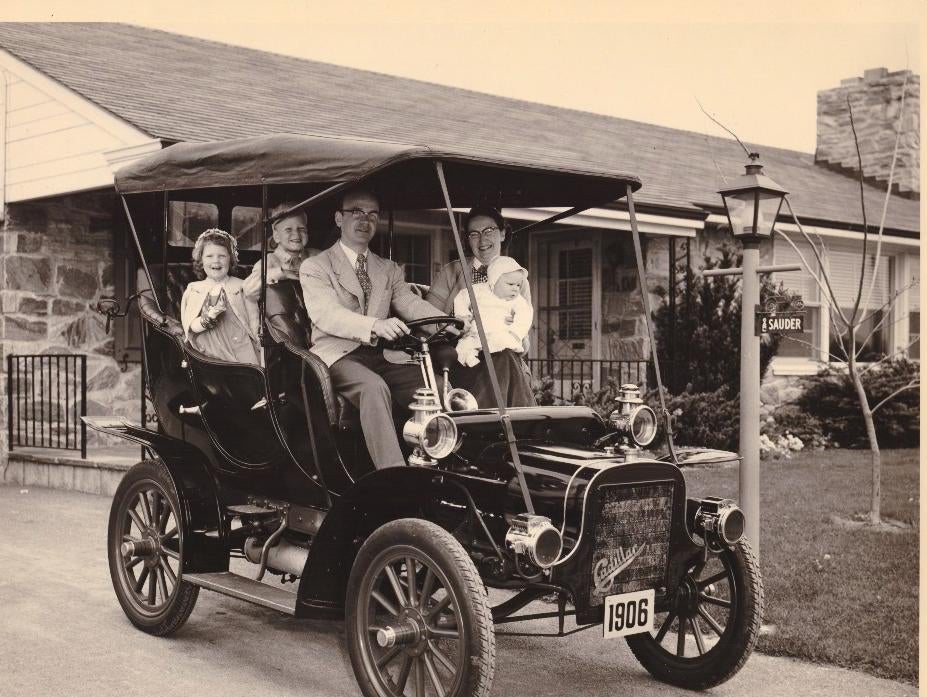 A vintage 1906 Cadillac car with a family dressed in early 20th-century attire, sitting inside and smiling in front of a house