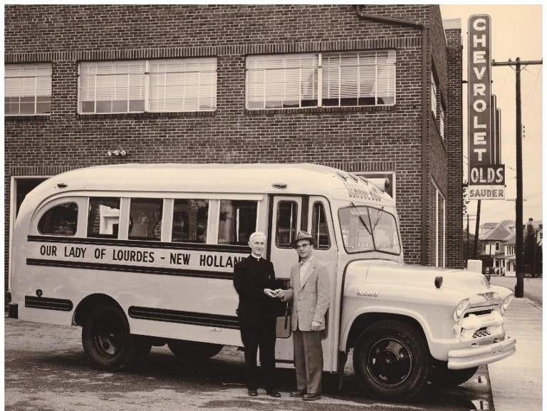 An old Chevrolet dealership building with a vintage bus labeled 'Our Lady of Lourdes - New Holland' parked in front, along with two men standing beside it