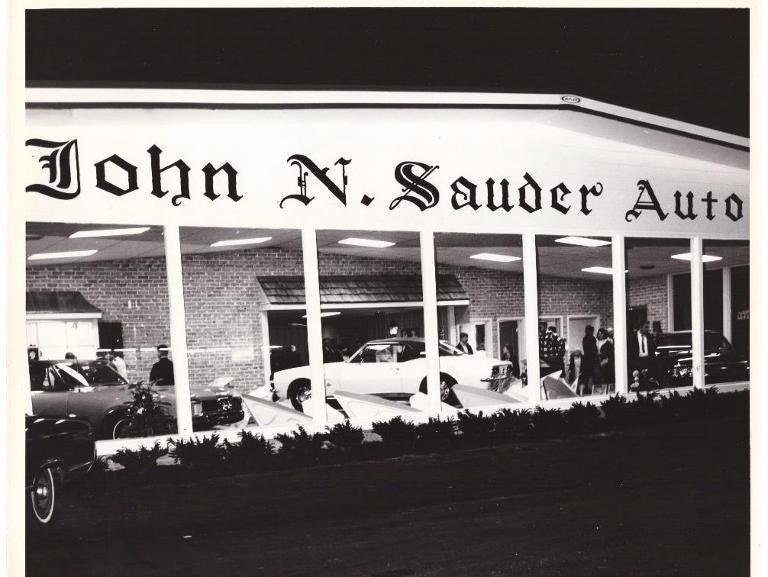 A historical black and white photograph of the John N. Sauder Auto dealership with classic cars parked in front